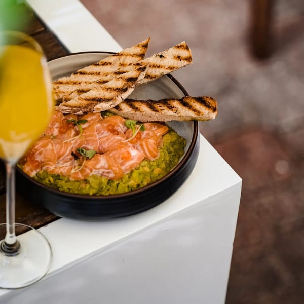 Bowl of green dip topped with sliced salmon, grilled flatbread, and microgreens beside a yellow drink.