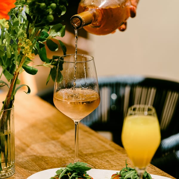 Rosé wine being poured into a glass on a wooden table with food and flowers.