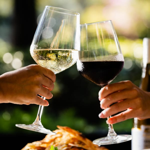 Two hands clinking wine glasses, one with white wine and one with red wine, outdoors.