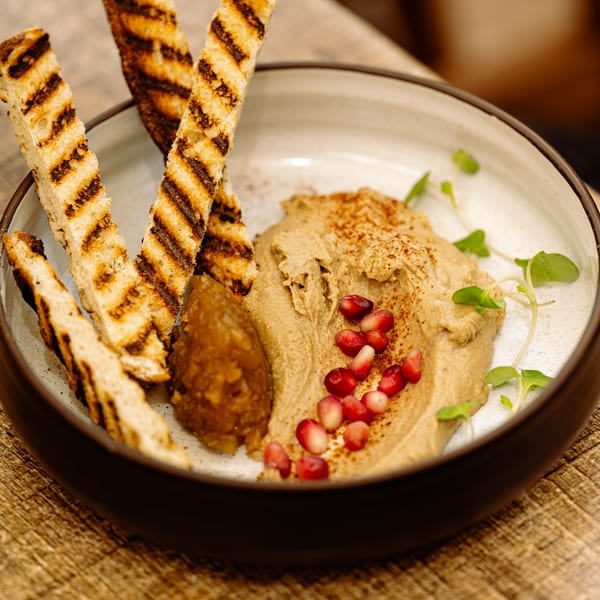 A bowl of hummus with pomegranate seeds, microgreens, and grilled bread on a wooden table.