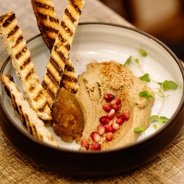 A bowl of hummus with pomegranate seeds, microgreens, and a side of grilled bread.
