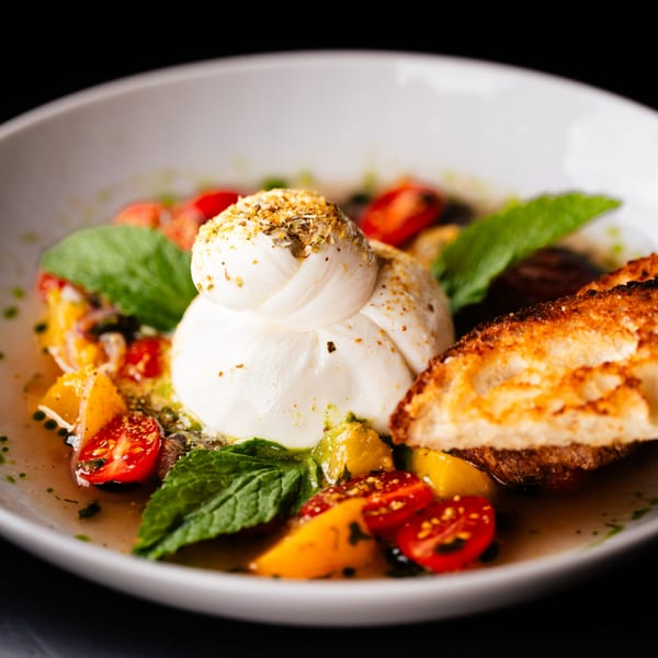 Burrata cheese with cherry tomatoes, mint, and toasted bread in a white bowl.