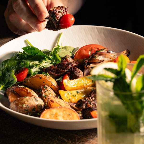 A hand holds a skewer with steak and cherry tomato over a bowl of steak, potatoes, and salad.