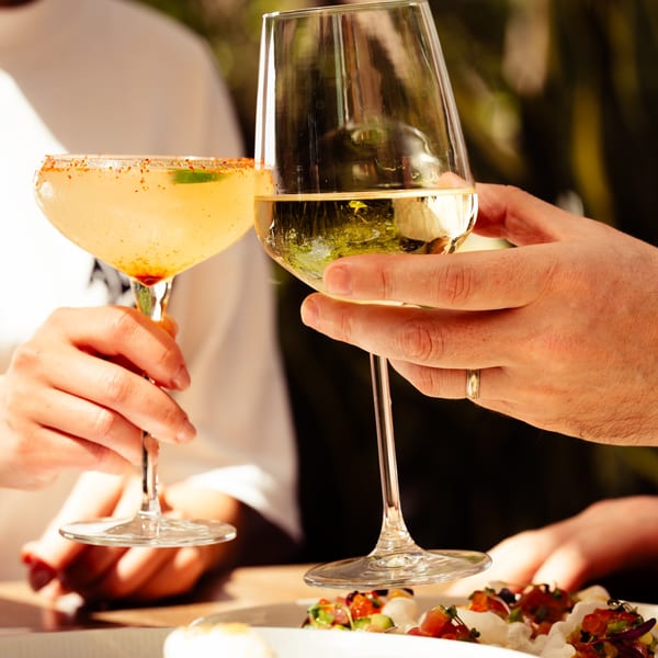 Two people clinking glasses, one with a margarita and the other with white wine, over a dish.