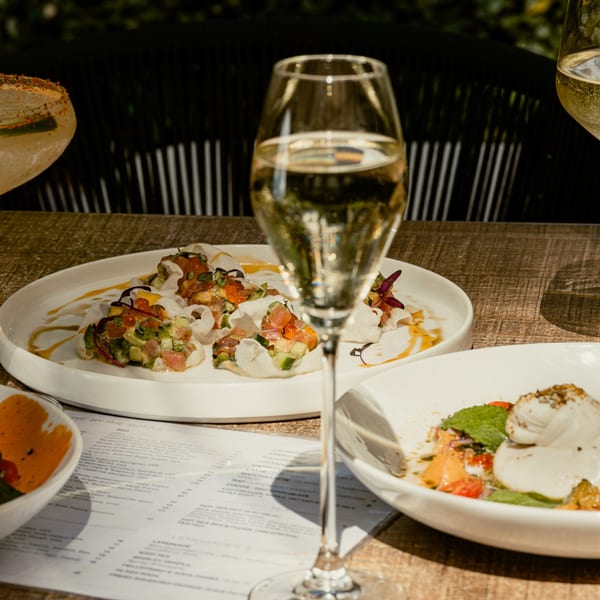 A glass of champagne in the foreground, with plates of appetizers and a menu on a wooden table.