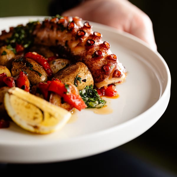 Grilled octopus with roasted potatoes, red peppers, herbs, and a lemon wedge on a white plate.