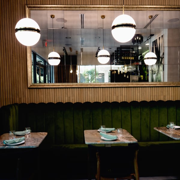 A restaurant interior with green velvet booths, marble tables, and spherical pendant lights reflected in a mirror.