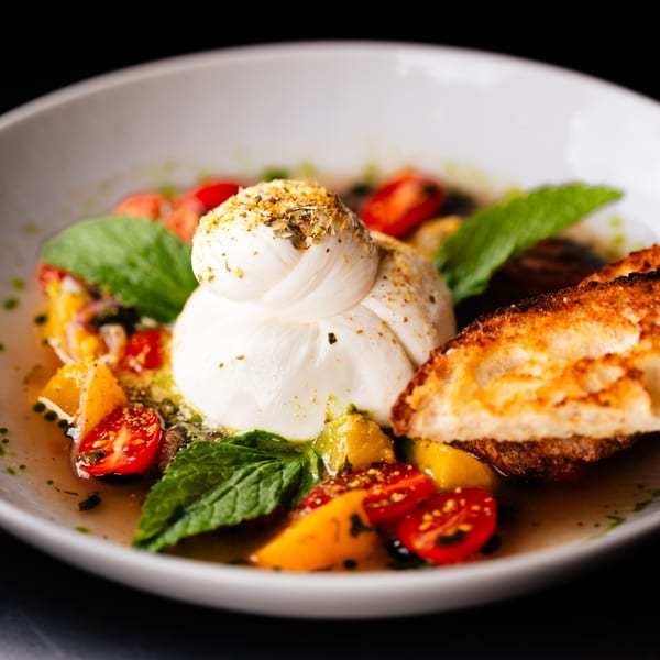 Burrata cheese with cherry tomatoes, mint, and toasted bread in a white bowl.