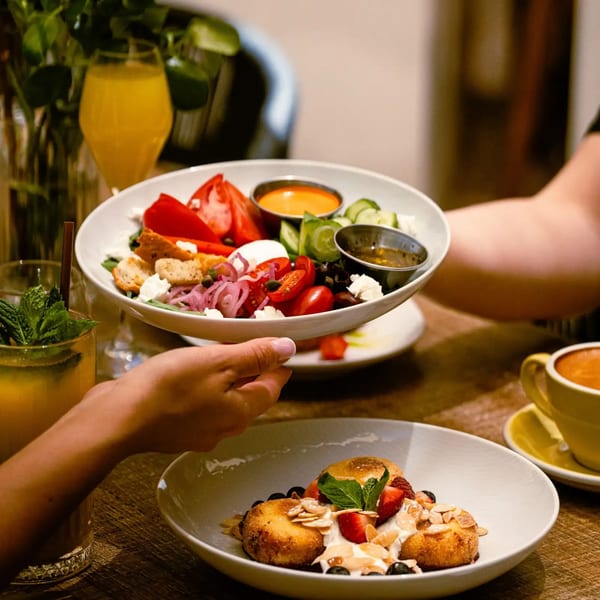 Hands serve a colorful Mediterranean salad with vegetables, feta, and sauces alongside a dessert garnished with berries and mint.