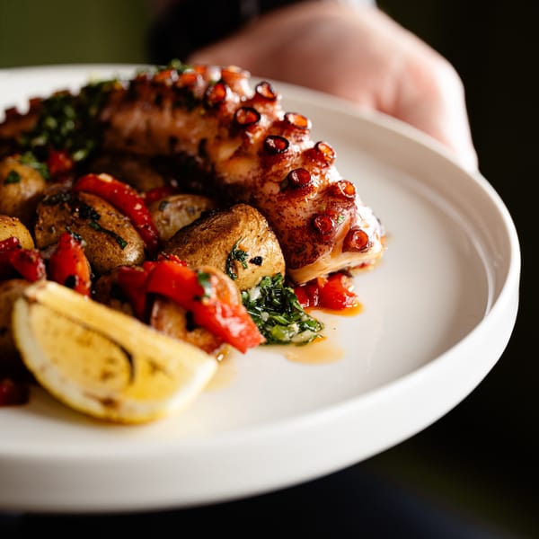 Grilled octopus with roasted potatoes, red peppers, and a lemon wedge on a white plate.