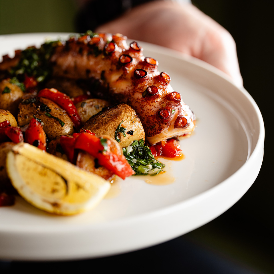 Grilled octopus with roasted potatoes, red peppers, and a lemon wedge on a white plate.